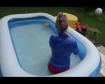 Mara playing with water in a small swimmingpool wearing a red shiny nylon shorts and a rain jacket (Video)