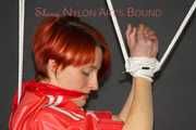 Sonja wearing a black rain pants and a red shiny nylon rain jacket tied and gagged with ropes overhead (Pics)