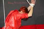 Sonja wearing a black rain pants and a red shiny nylon rain jacket tied and gagged with ropes overhead (Pics)