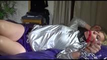 Pia tied and gagged on bed with cuffs and neckband wearing a sexy purple shiny nylon shorts and a silver rain jacket (Video)